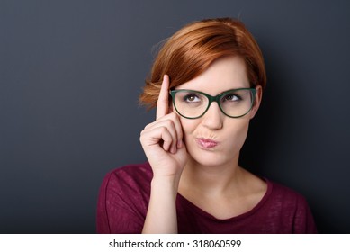 Nerdy scholastic young woman wearing geeky glasses standing thinking with her finger raised and a grimace of concentration in a humorous stereotypical depiction, over a dark background with copyspace