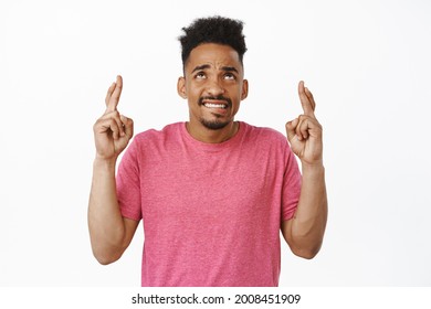 Nervous african american guy praying god, looking up worried with fingers crossed, making wish, anticipating important news with scrared anxious face, standing over white background