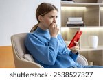 Nervous anxious woman receives unpleasant message on phone frowning, thinking about troubles, worried bites fingernails at home. Exhausted with thoughts female sitting with smartphone feeling stress.