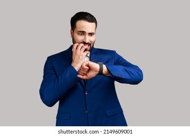 Nervous bearded man biting finger nails and looking at his wrist clock with anxious, worried about deadline, wearing official style suit. Indoor studio shot isolated on gray background.