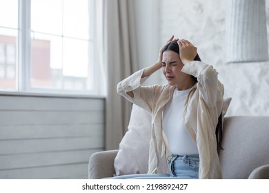 Nervous breakdown, depression, panic attack. A young beautiful brunette woman sits on the sofa at home and holds her head with her hands, is nervous, feels pain, discomfort.