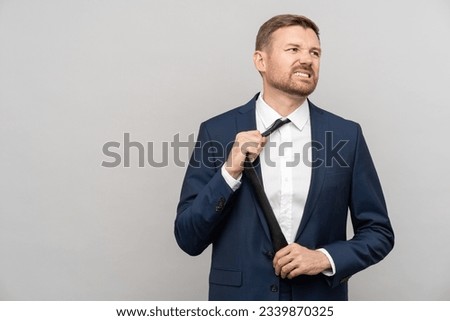 Nervous businessman putting off tie wearing in official blue suit with dissatisfied face isolated on grey studio background. Discomfort, unsuitable clothes. Annoyed office worker annoyed by dress code