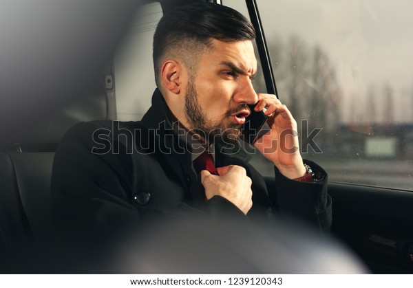Nervous businessman sitting\
in luxury car. Young bearded man wearing formal attire talking by\
smartphone