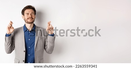 Nervous businessman in suit begging god, holding fingers crossed with eyes closed, pleading to win, making wish on white background.