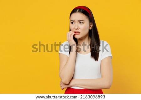 Nervous disturb young sad woman of Asian ethnicity 20s years old wears white t-shirt looking aside biting nails isolated on plain yellow background studio portrait. People emotions lifestyle concept