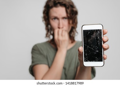 Nervous Embarassed Girl Bites Nails, Holding Her Broken Smartphone, Isolated On Gray Background.