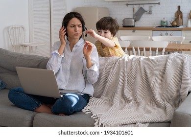 Nervous Freelancer Mother Sitting On Couch At Home Office During Lockdown, Working On Laptop. Little Child Distracts From Work, Taking Off Headphones, Making Noise And Asking Attention From Busy Mom