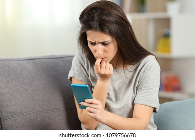 Nervous girl biting nails reading phone content sitting on a couch in the living room at home
