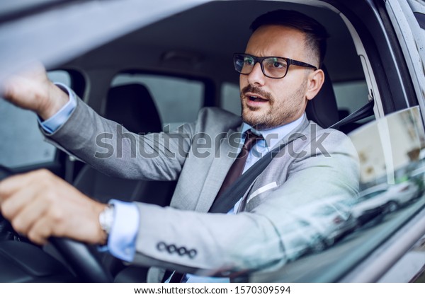 Nervous handsome caucasian businessman in suit and\
with eyeglasses commenting traffic while sitting in his car. Hand\
is on steering wheel.