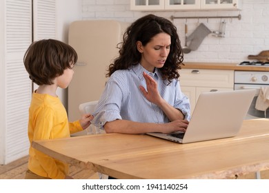 Nervous Mom Sits By The Table At Home Office During Lockdown, Working On Laptop, Asks Not To Make Noise. Noisy Child Distracts From Work, Pulls The Sleeve, Asking Attention From Annoyed Busy Mother. 