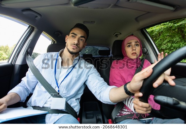 Nervous Muslim Lady In Hijab Having Driving\
Lesson With Instructor, Male Teacher Helping Islamic Female Student\
To Drive Car, Holding Steering Wheel, Closeup Shot Inside Of\
Automobile Vehicle