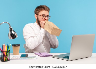 Nervous overemotional man office worker deeply inhaling and exhaling holding paper breathing bag near mouth, sitting at workplace with laptop. Indoor studio shot isolated on blue background