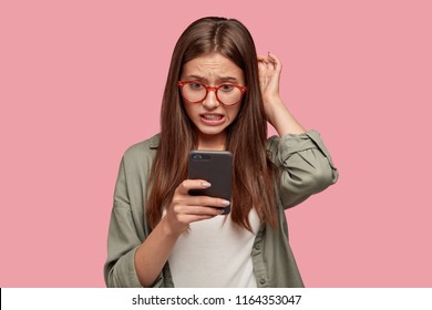 Nervous puzzled woman scratches head in bewilderment, reads bad news at internet, uses modern cell phone, wears fashionable clothes, worries aboutt something, stands in studio against pink wall - Powered by Shutterstock