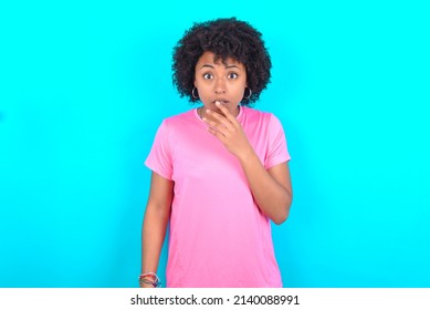 Nervous puzzled young girl with afro hair style wearing sport pink t-shirt over blue background opens mouth from surprise, reacts on sudden news.
