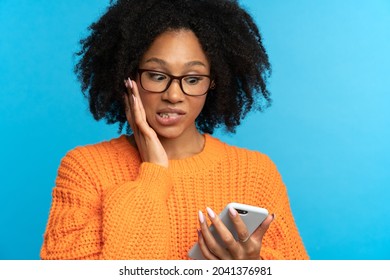 Nervous Shocked African Ethnicity Woman Looking At Smartphone Bites Lips. Mixed Race Young Girl Reading Bad News, Notification Unpleasant Message On Cellphone Isolated On Studio Blue Color Background