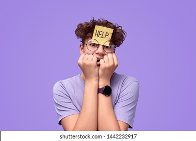 Nervous Teen Boy With Sticky Note On Forehead Biting Nails And Looking At Camera While Asking For Help Before Exam Against Violet Background