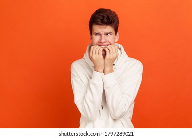 Nervous terrified teenager in white casual style sweatshirt biting his fingers with shocked look, fears and phobias. Indoor studio shot isolated on orange background