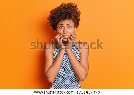 Nervous worried Afro American woman clenches teeth looks embarrassed at camera wears fashiobnable outfit afraids of bad consquences poses in studio against orange background. Scared female model