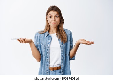 Nervous Young Woman Shrugging With Hands Spread Sideways, Holding Phone And Frowning Troubled, Having Problem On Smartphone, Standing Over White Background