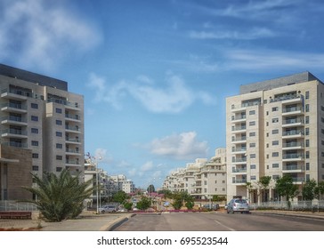 Nes Ziona, Israel-March 20, 2016: Front View Of Two Modern 8 Story Residential Condominiums Are Located On Both Sides Of The Beginning Of Khoshen Street. Wide Urban Road And Blue Sky. Horizontal Shot