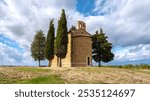 Nestled among vibrant green fields, a quaint chapel stands tall, embraced by majestic cypress trees. The sky displays rich hues as clouds drift lazily above, capturing the tranquil beauty of Tuscany.