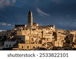 Nestled in the hills of Puglia, Matera reveals its ancient cave dwellings as golden light bathes the historic architecture, creating a striking contrast against the stormy sky at sunset.