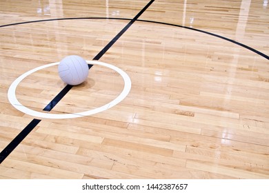 A Netball Ball placed within the centre circle on an indoor Netball court, positioned left of shot. The indoor wooden floorboards are a light coloured grain reflecting the lights from above.