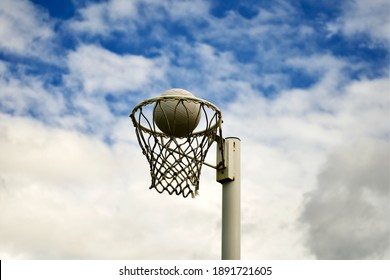 A Netball Positioned Just Inside The Ring Of A Netball Goal On A Cloudy Day.