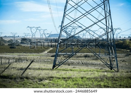 A network of high voltage power lines stretches across a rural landscape, with tall metal towers supporting the electrical cables. 