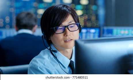 In The Network Operations Center Senior Trader Teaches Young Associate To Makes Personal Client Call With A Headset. They're Surrounded By Monitors Showing Data And Graphs.