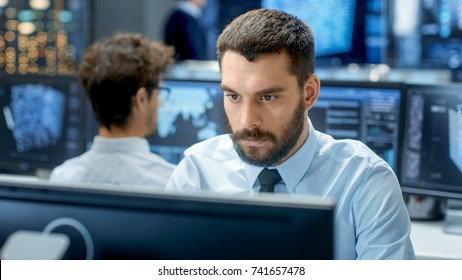 Neural Network And Machine Learning Engineer Programming At His Workstation. Office Is Crowded With People Working. Multiple Displays Show Neural Network, Artificial Intelligence Representation.