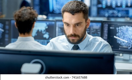 Neural Network And Machine Learning Engineer Programming At His Workstation. Office Is Crowded With People Working. Multiple Displays Show Neural Network, Artificial Intelligence Representation.