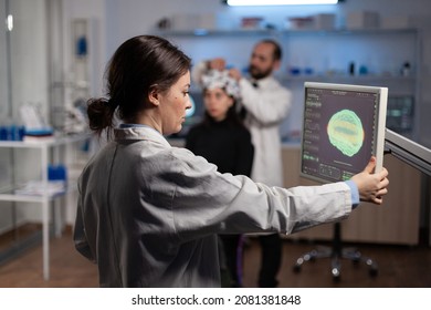 Neurologist woman doctor looking at monitor with medical tomography analyzing brain activity using high tech equipment. In background specialist man adjusting eeg headset of woman patient - Powered by Shutterstock