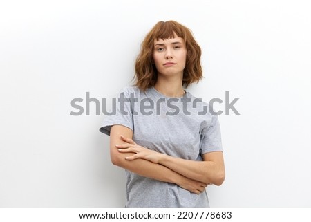 a neutral, emotionless woman with straight hair stands against a light background in a gray cotton T-shirt and looks at the camera with her arms crossed on her chest Stock photo © 