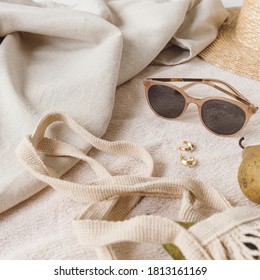 Neutral fashion composition with women's accessories and bijouterie on beige blanket. String bag, straw hat, sunglasses, rings, earrings, pear. Minimal lifestyle concept.