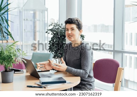 Neutral gender middle aged woman in casual clothing having conversation in office while working laptop on workstation in open space office, coworking, Remote workplace, colaboration, selective focus
