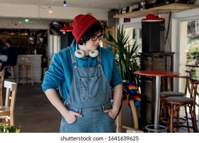 Neutral gender teenager wearing red hat, headphones and denim overalls in a modern cafe - Powered by Shutterstock