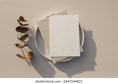 Neutral wedding stationery. Blank greeting card, envelope mockup in sunlight. Invitation, template on plate. Dry eucalyptus tree branch. Table background, shadows, floral composition. Flatlay, top 