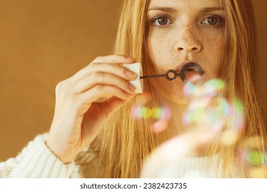 Never get old concept. Funny portrait of fashion model with long red hair in white sweater blowing party bubbles over wooden background. Daylight. Close up. Text space. Studio shot