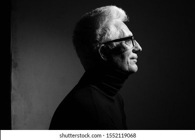Never get old, handsome at every age concept. Close up portrait of fashionable mature man wearing trendy eyewear, black turtleneck, sitting in art gallery. Modern haircut. Silver hair. Graphic shadows