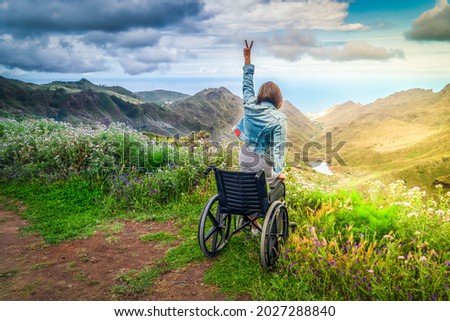Never give up. Rear view of young handicapped woman sitting on wheelchair on top of mountain with hands up and looking at amazing nature landscape while traveling alone. International Disability Day