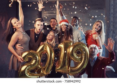 New 2019 Year is coming! Group of cheerful young multiethnic people in Santa hats carrying gold colored numbers and throwing confetti on the party.