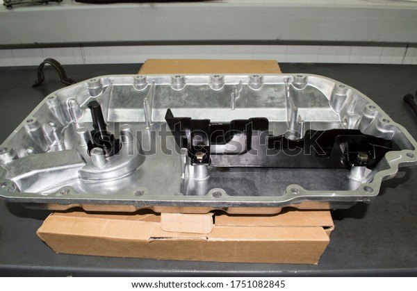 The new aluminum\
oil pan of the internal combustion engine sits on a cardboard box\
on a gray metal work table