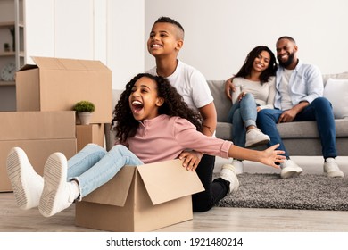 At New Apartment. Happy African American Family Having Fun And Celebrating Moving Day, Excited Brother Riding His Sister In Cardboard Box, Parents Hugging While Sitting On The Couch In Background