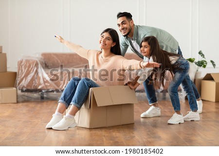 At New Apartment. Happy loving family having fun and celebrating moving day, excited man and little girl riding smiling woman in cardboard box, fooling around in new flat in living room