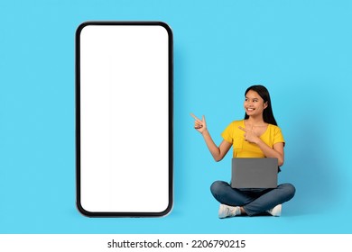 New App For Work And Study. Happy Young Chinese Lady Sit With Laptop And Pointing Fingers At Smartphone With Blank Screen, Isolated On Blue Background, Studio. Recommendation And Advice, Ad And Offer