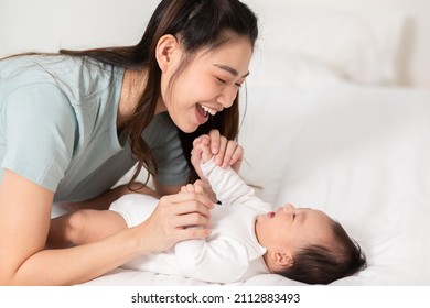 New asian mom playing to adorable newborn baby on bed smiling and happiness at home.Mom talking with infant baby and holding her hands laughing together.Baby and Mother day concept