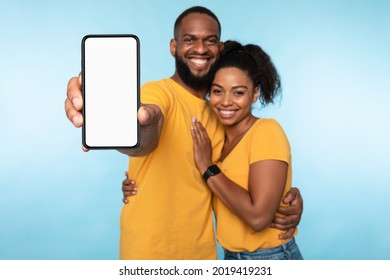 New awesome mobile app. African American couple presenting smartphone with mockup, promoting application or website, advertising your product or service, blue studio background. Selective focus