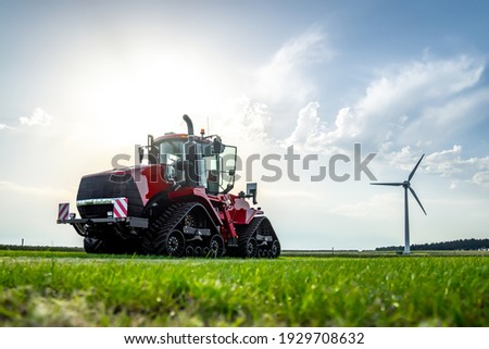 New big red modern farm tractor with quad tracks for powerful combine with renewable energy food and power crisis parked sunshine farmers rural agricultural field with wind turbine electric generator.