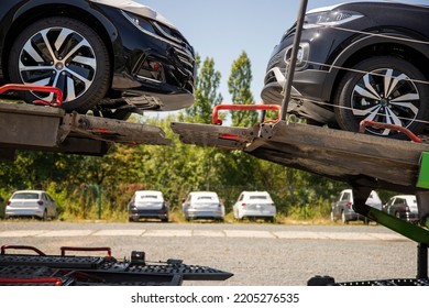 New Black Cars - 4-door Coupé And Crossover - On Car-carrying Trailer, Car Hauler, Auto Transport Trailer, Semi-trailer. Visible Headlights. Side View, Other Cars And Trees In The Background.